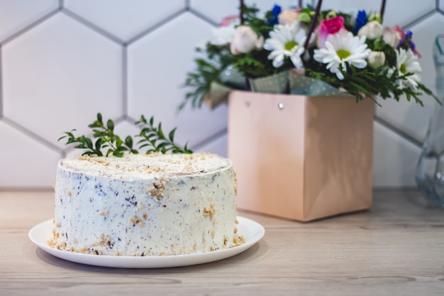 Eenvoudige zelfgemaakte witte cake met noten op het aanrecht in de keuken op de achtergrond van een boeket van verschillende bloemen in een papieren zak met handvatten