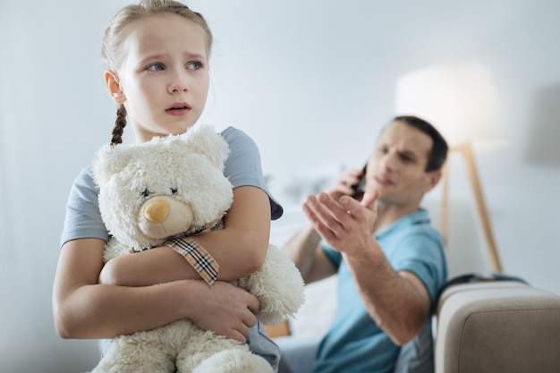 Eenzaam blauwogig blond meisje dat haar speeltje vasthoudt en huilt terwijl haar vader aan de telefoon zit