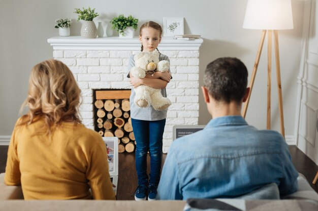 Foto eenzaam blond meisje dat haar teddybeer vasthoudt en voor haar ouders staat terwijl ze op hun laptops werken