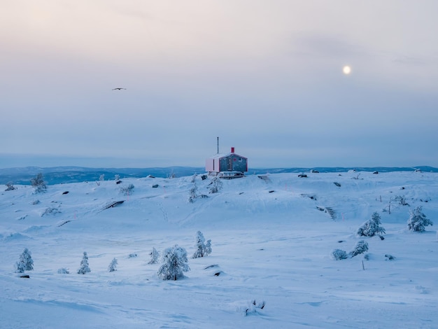 Eenzame rode bungalow op een besneeuwde, ijzige heuvel onder een donkere avondhemel met een volle maan minimalistische achtergrond met een eenzame rode hut in de winter