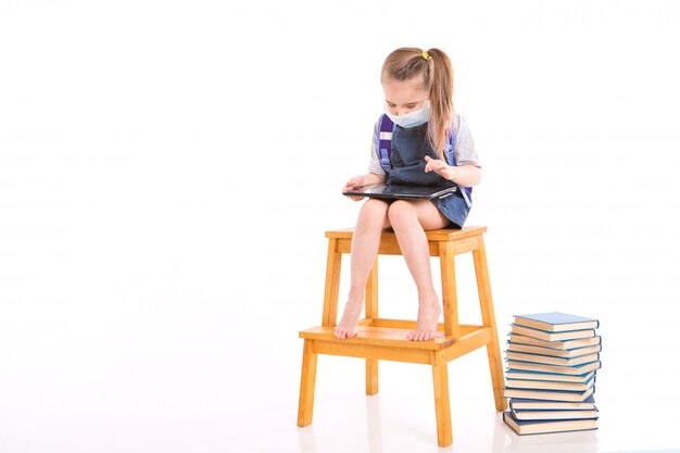 eerste-klasser in medisch masker leert lezen. klein meisje op thuis afstandsonderwijs. kind in medische masker werkt op laptop en boeken. huiswerk aan het maken.