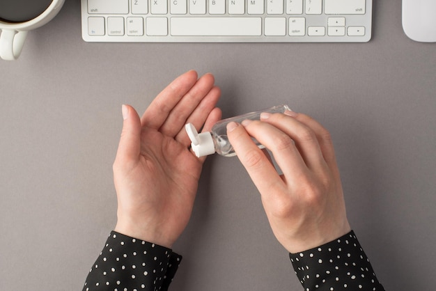 Eerste persoon bovenaanzicht foto van de handen van de vrouw met behulp van ontsmettingsmiddel transparante fles witte toetsenbordmuis en kopje koffie op geïsoleerde grijze achtergrond