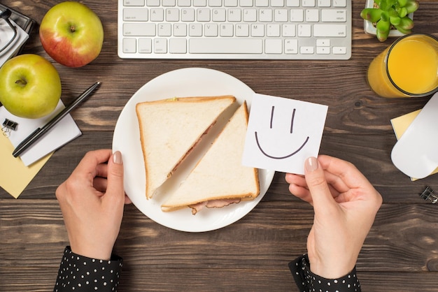 Eerste persoon bovenaanzicht foto van hand met sticker notitie met getekende lachende gezicht over bord met toast toetsenbord muis appels glas sap plant en briefpapier geïsoleerde donkere houten bureau achtergrond