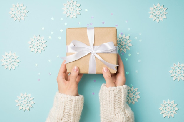 Eerste persoon bovenaanzicht foto van handen in witte trui met ambachtelijke papieren geschenkdoos met witte strik over grote decoratieve sneeuwvlokken en glanzende confetti op geïsoleerde pastelblauwe achtergrond