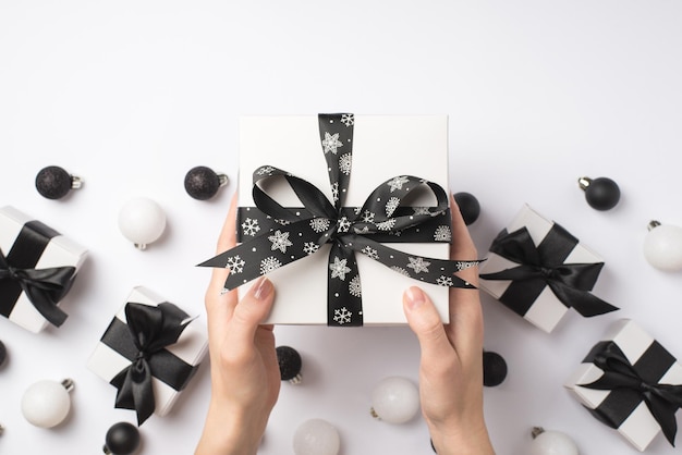 Eerste persoon bovenaanzicht foto van jonge meisje handen geven witte geschenkdoos met zwarte satijnen lint strik over presenteert witte en zwarte kerstboom ballen op geïsoleerde witte achtergrond met lege ruimte