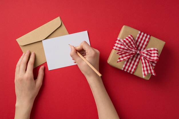 Eerste persoon bovenaanzicht foto van valentijnsdag decor vrouwelijke handen schrijven brief envelop en ambachtelijke papier geschenkdoos met geruite strik op geïsoleerde rode achtergrond met copyspace