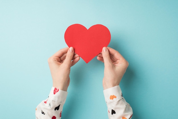 Eerste persoon bovenaanzicht foto van valentijnsdag decoraties vrouwelijke handen in wit overhemd met rood papieren hart op geïsoleerde pastelblauwe achtergrond met lege ruimte