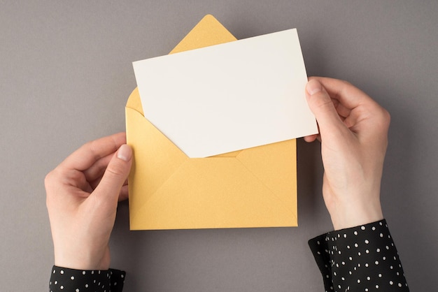 Eerste persoon bovenaanzicht foto van vrouwelijke handen met open pastelgele envelop met witte kaart op geïsoleerde grijze achtergrond met lege ruimte