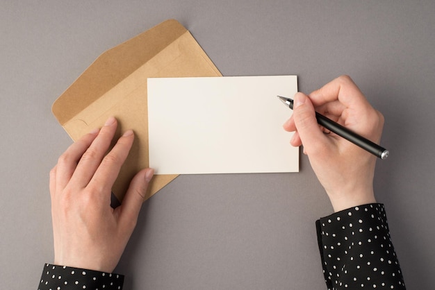 Eerste persoon bovenaanzicht foto van vrouwelijke handen met pen over witte kaart open ambachtelijke papieren envelop op geïsoleerde grijze achtergrond met lege ruimte