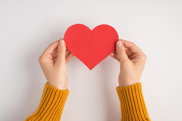 Eerste persoonsfoto van de handen van de vrouw in gele trui met rood papieren hart op een geïsoleerde witte achtergrond met lege ruimte