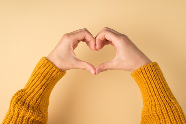 Foto eerste persoonsfoto van vrouwenhanden in gele trui die hart maken met vingers op geïsoleerde pasteloranje achtergrond