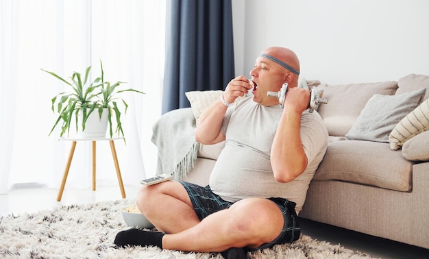 Eet en doet oefeningen Grappige dikke man in vrijetijdskleding is binnenshuis thuis