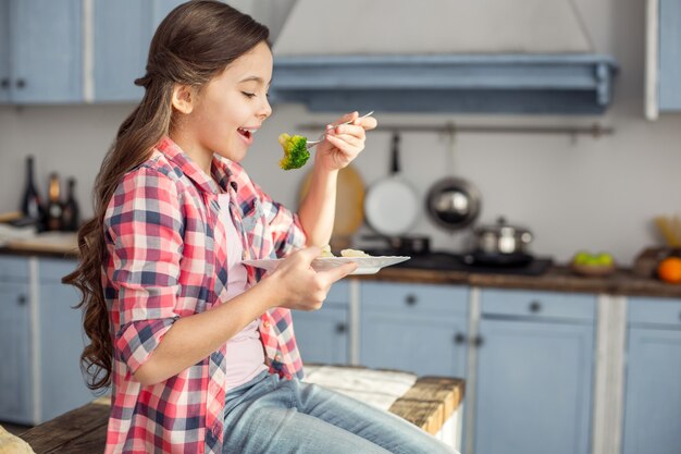 Eet smakelijk. Mooie vrolijke donkerharige meisje glimlachend en zittend op de tafel en groenten eten voor het ontbijt