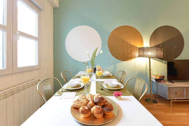 Eetkamer met rechthoekige witte houten tafel, metalen stoelen, muffins en gebak voor het ontbijt en vers geperst sinaasappelsap