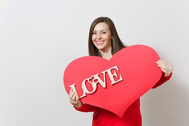 Effective young smiling woman in red casual clothes holding big red heart, wooden word love on white background. Copy space for advertisement. St. Valentine's Day or International Women's Day concept.