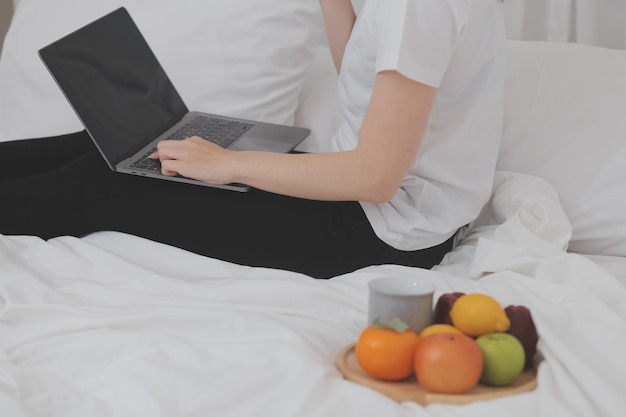 Efficiënt jong millennial meisje dat in de ochtend op een bed zit, een laptop gebruikt en croissants eet en koffie drinkt voor het ontbijt