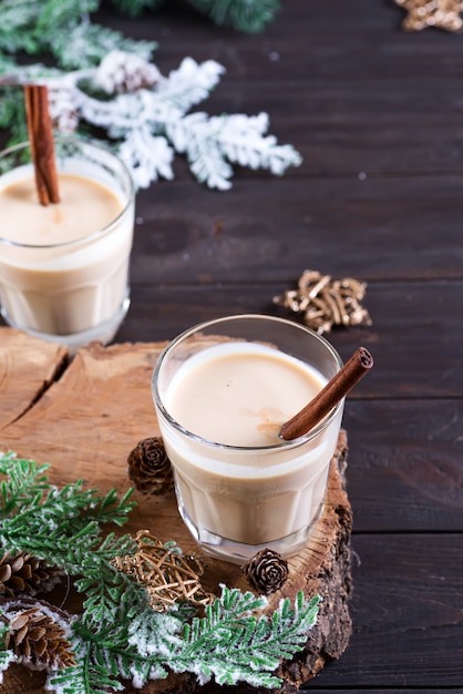 Eggnog Christmas milk cocktail with cinnamon, served in two glasses on a wood cutting board with fir branch and pine cone on dark wooden 