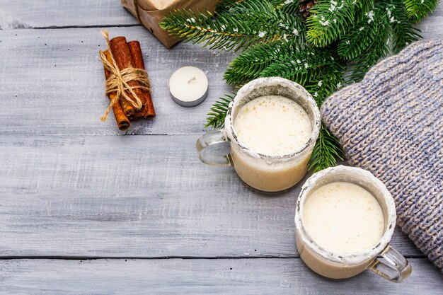 Eggnog коктейль в кружку двух стаканов, аранжировано с рождественские украшения на светлом деревянном столе. вечнозеленая еловая ветка, подарок, уютный трикотаж, искусственный снег