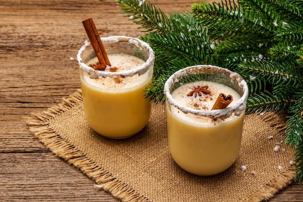 Eggnog cocktail in two glasses arranged with christmas decoration on old wooden table. Evergreen fer tree branch, artificial snow, sackcloth napkin