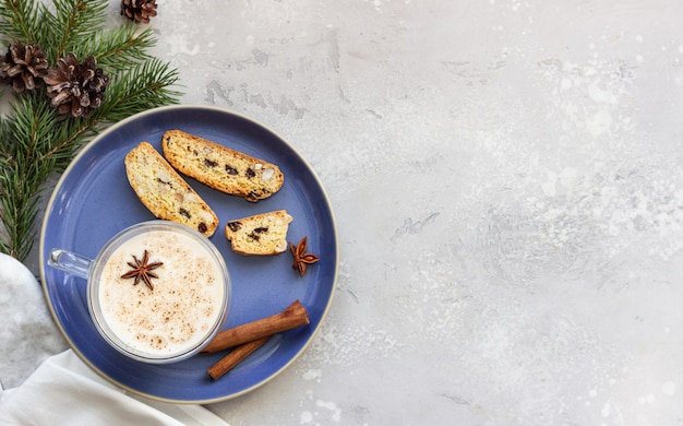 Eggnog served in glass mug with biscotti, winter spices, fir branches and cones.