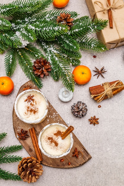 Eggnog with cinnamon and nutmeg for Christmas and winter holidays. Homemade beverage in glasses with spicy rim. Tangerines, candles, gift. 