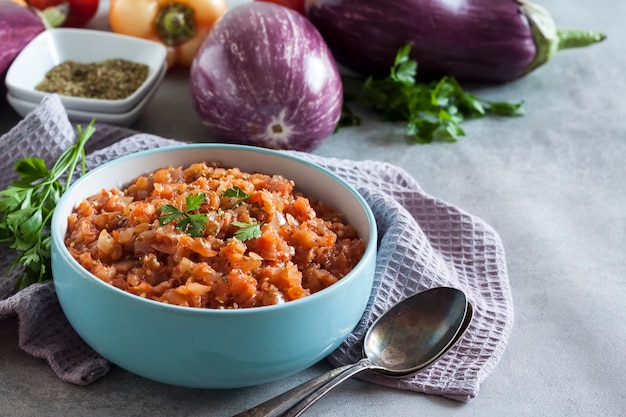 Eggplant caviar in blue bowl and fresh vegetables on background.