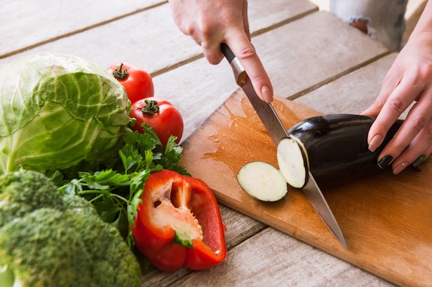 Eggplant cutting on wooden desk by unrecognizable woman. Red pepper, broccoli, tomatoes, cabbage and parsley included, top view picture of cooking process