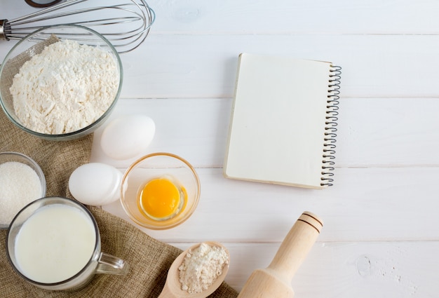 Eggs, flour, milk, with whisk on white wooden table from above.