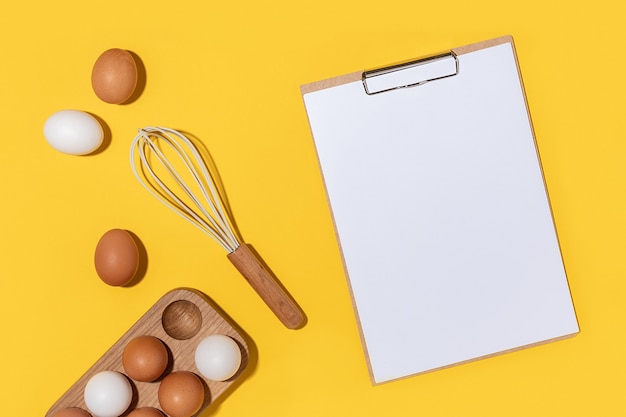 Eggs in wooden egg box, whisk and clipboard with white paper on yellow background. Flat lay Top view Mockup.