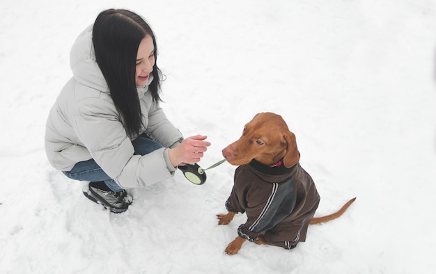 Eigenaar en een schattige bruine hond in de kleren zitten in de sneeuw en spelen.