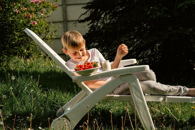 An eightyearold boy eats strawberries with yogurt in the summer in the yard