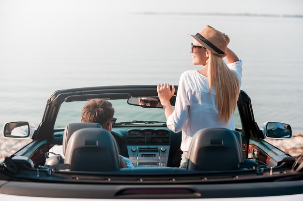 Eindelijk aangekomen! Achteraanzicht van een lachende jonge vrouw die van het landschap geniet terwijl haar vriendje op de voorstoel van hun cabrio zit