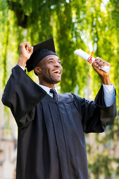 Eindelijk geslaagd! gelukkige jonge afrikaanse man in afstudeerjurken met diploma en stijgende armen omhoog