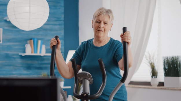 고정식 자전거에서 사이클링 운동을 하는 노인. 자전거로 사이클 활동을 하기 위해 정적 유산소 기계로 훈련하는 나이든 여성. 피트니스 및 운동 장비를 사용하는 노인