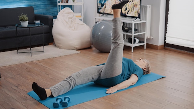 Elder adult raising legs to do physical exercise and workout on yoga mat. Senior person doing muscles stretching activity and gymnastics training at home. Fitness exercising for strength