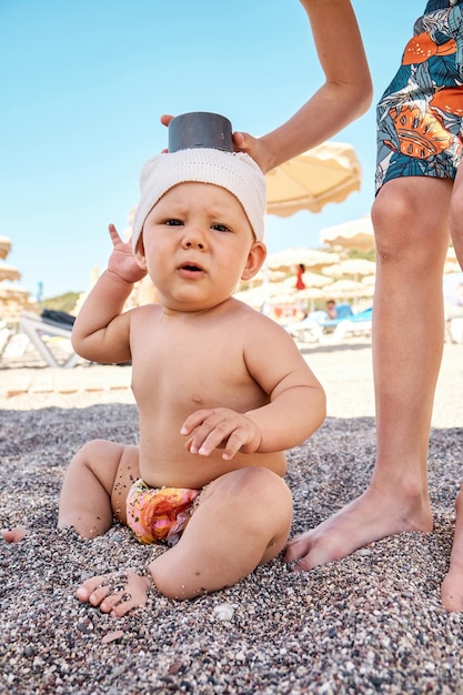 Elder brother plays with baby boy sibling sitting on sand in shadow from umbrella