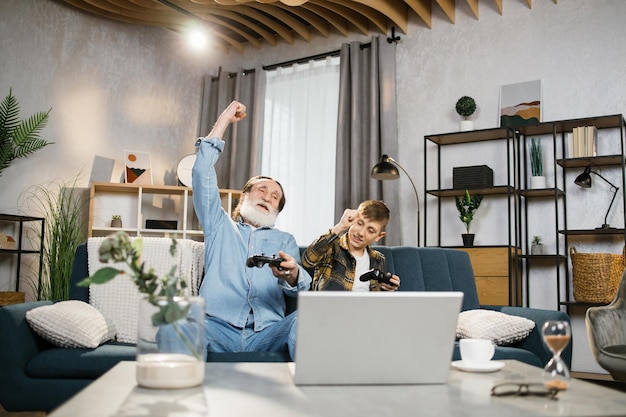 Elder man and his grandson sitting on couch and using wireless joysticks for playing video games