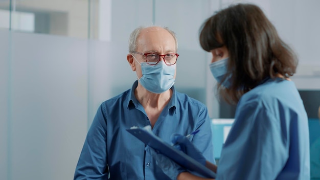 Elder patient attending checkup examination with nurse in\
office, talking about alternative medicine and treatment. health\
assistant taking notes and discussing with old man during\
pandemic.