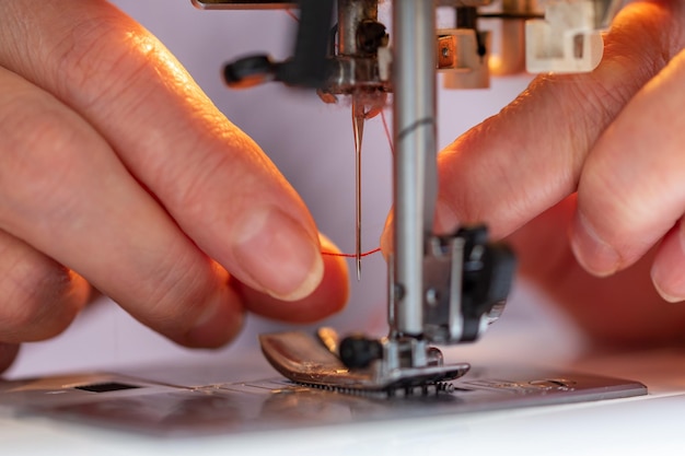 Elder woman fingers inserting a red thread into a sewing machine's needle closeup