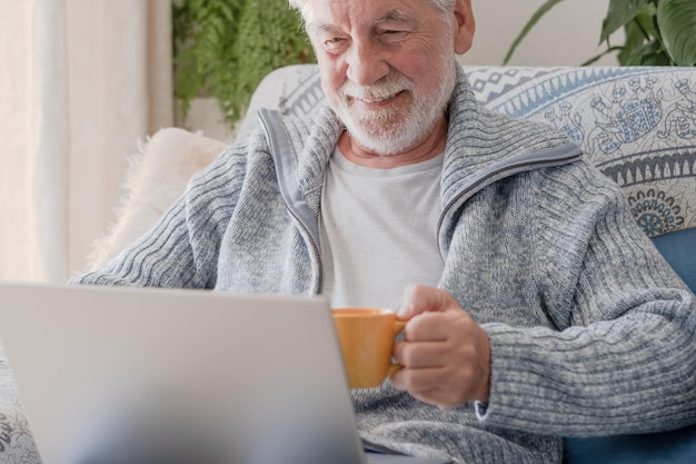 Elderly 70s man seated on sofa browsing on laptop senior looking at laptop screen surfing the net older generation and modern tech application easy usage concept