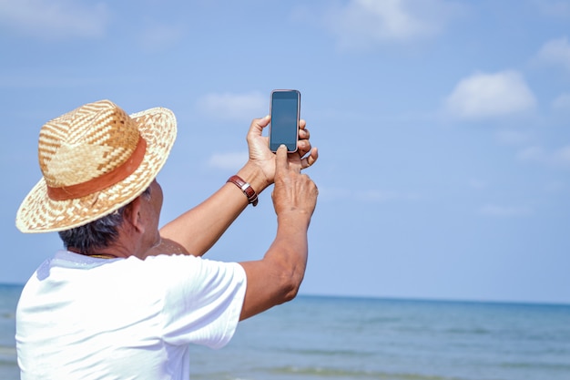 An elderly Asian man holding a phone to take pictures in the sea