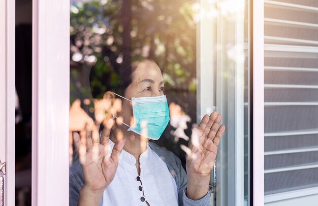 Elderly asian women wearing face mask with hands touching on window,stay at home during coronavirus and covid-19 epidemic or pandemic,social distancing concept