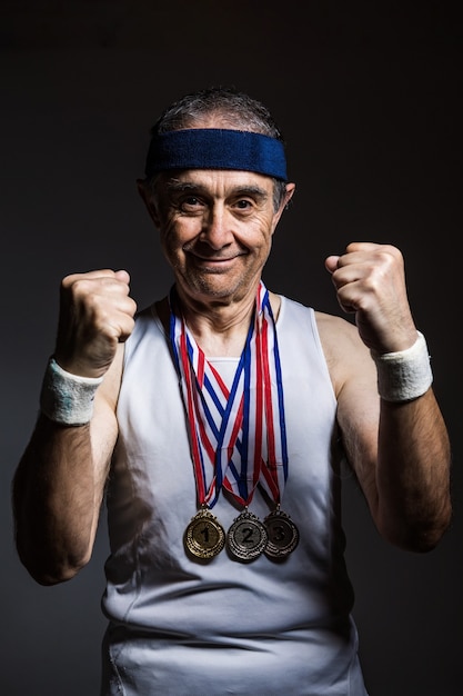 Elderly athlete wearing white sleeveless shirt, with sun marks on his arms, with three medals on his neck, clenching his fists, on a dark background. Sports and victory concept.