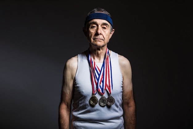 Пожилой спортсмен в белой майке, с солнечными отметинами на руках, с тремя медалями на шее, демонстрирующими их, на темном фоне. Концепция спорта и победы