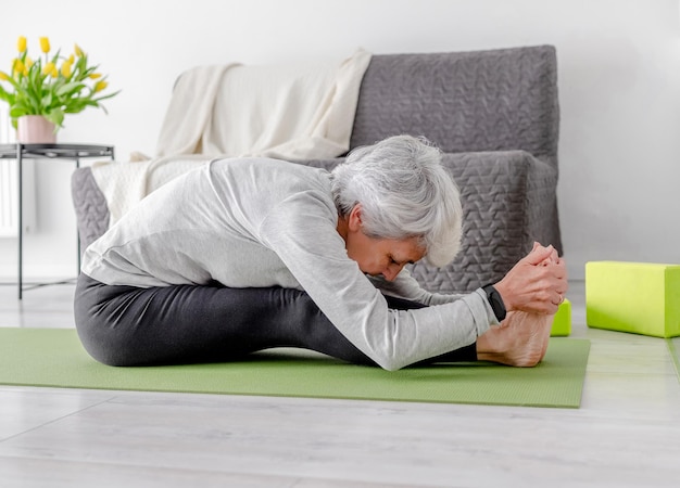 Elderly athletic woman bends while sitting on yoga mat at home on floor in bright room with flowers