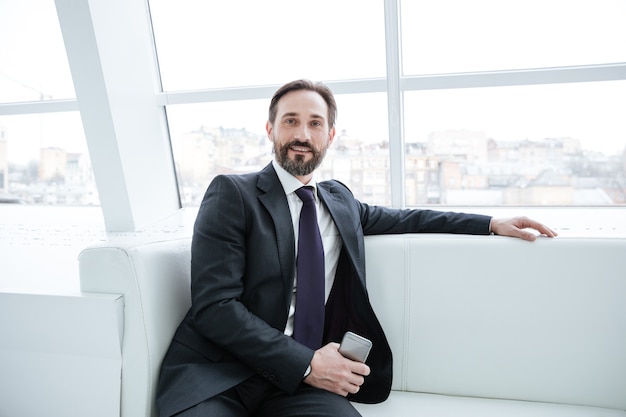 Elderly bearded business man sitting on sofa with phone near the window in office