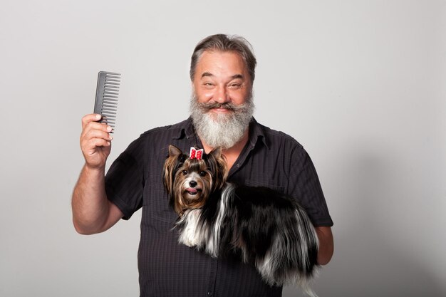 An elderly bearded hairdresser for dogs with a comb and a terrier on a gray background in the studio