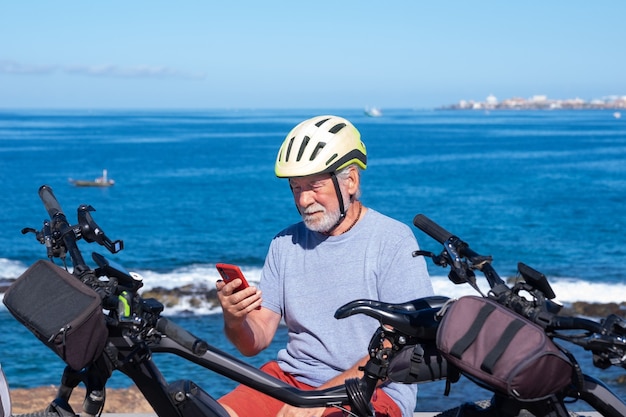 Elderly bearded man sitting in outdoor sea excursion sitting and using mobile phone. He wears bike helmet near the bicycle, horizon over sea. Active retiree uses technology