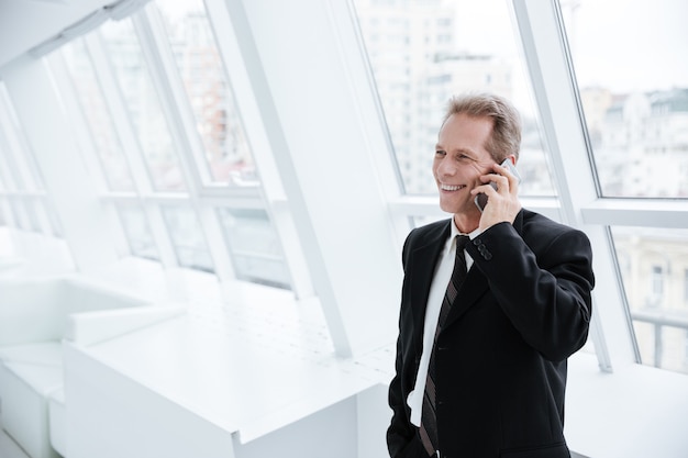 Elderly business man talking at phone and standing near the window in conference room