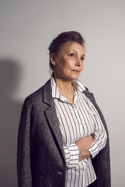 Elderly businessman woman in a white shirt and a gray jacket stands against a white wall